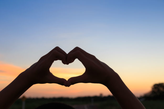 Female Hands In The Form Of Heart Against Sunlight In Sunset Sky